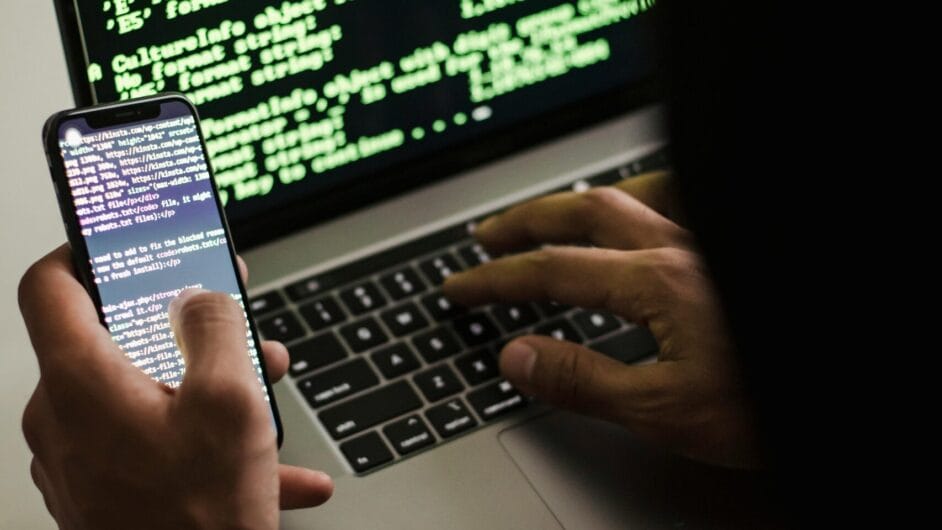 A hooded figure engaged in hacking using a laptop and smartphone in low light.